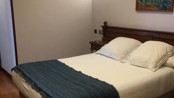Een net opgemaakt bed met witte lakens en een blauwe deken, een houten hoofdbord, een nachtkastje met een lamp en een schilderij aan de muur.