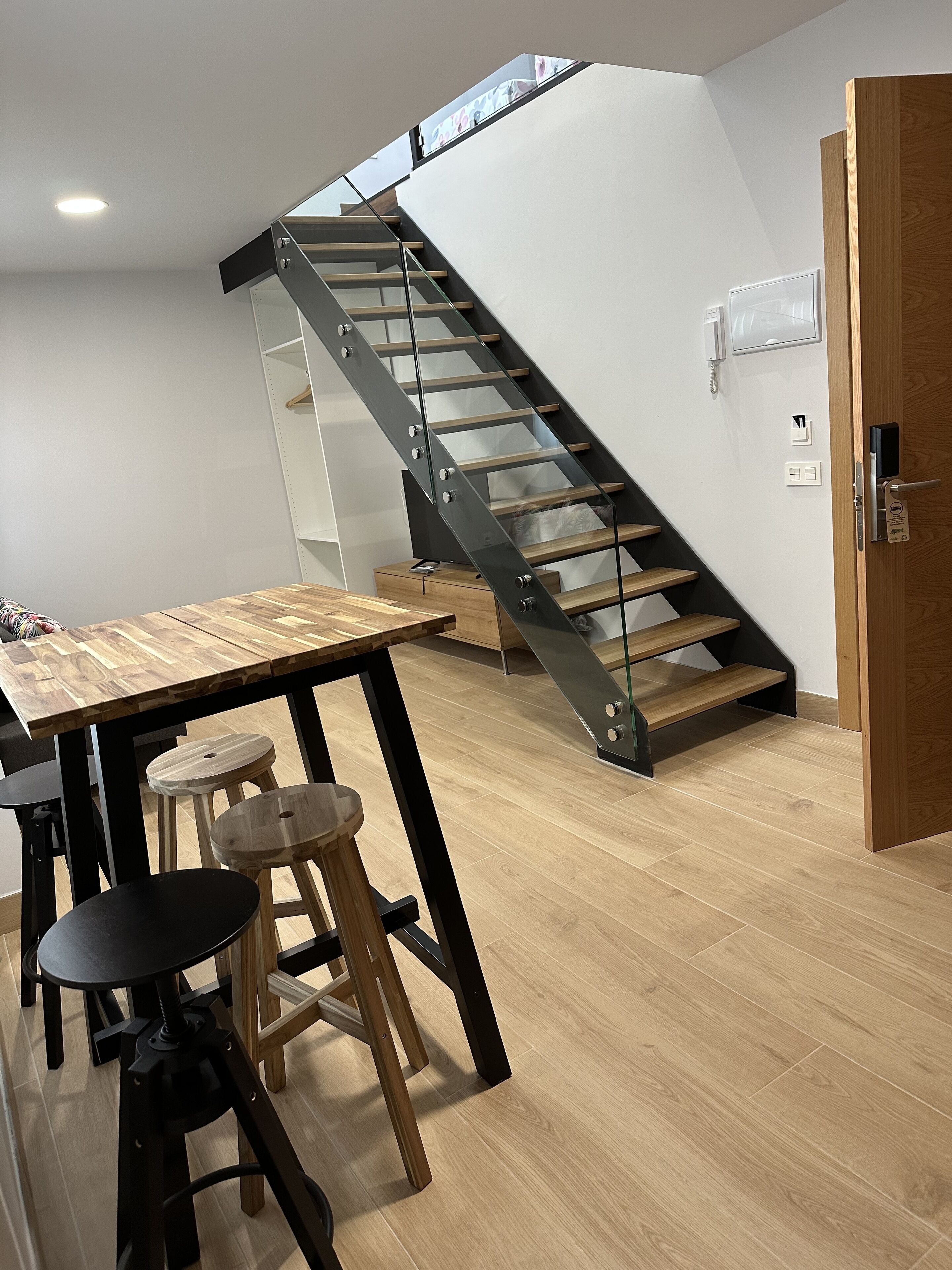 Un intérieur moderne avec une table à manger en bois, des tabourets, un escalier doté de rampes en verre et une porte en bois.