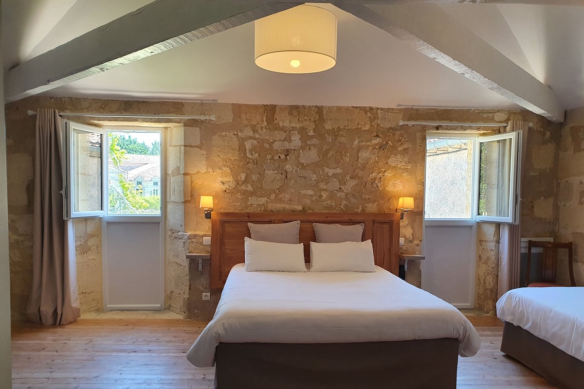 A bedroom with a stone wall, wooden ceiling beams, a large bed with a wooden headboard, two bedside lamps, and a window with curtains.