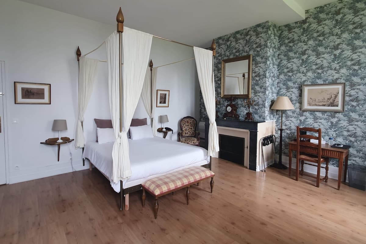 A bedroom with a four-poster bed, a desk with a chair, a fireplace, and a mirror on the wall.