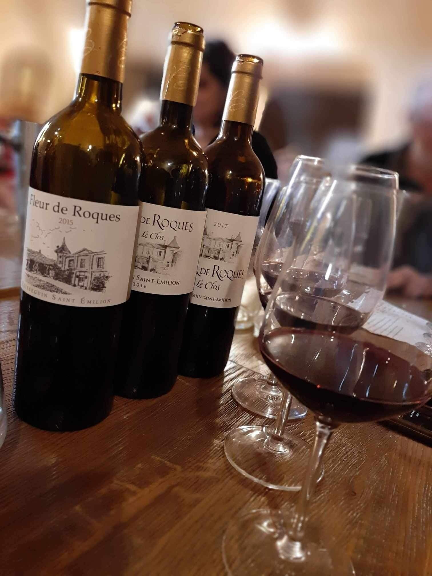 Trois bouteilles de vin portant l’étiquette « Fleur de Roques », avec les millésimes 2015 et 2017, et un verre à vin rempli de vin rouge, posés sur une table en bois.