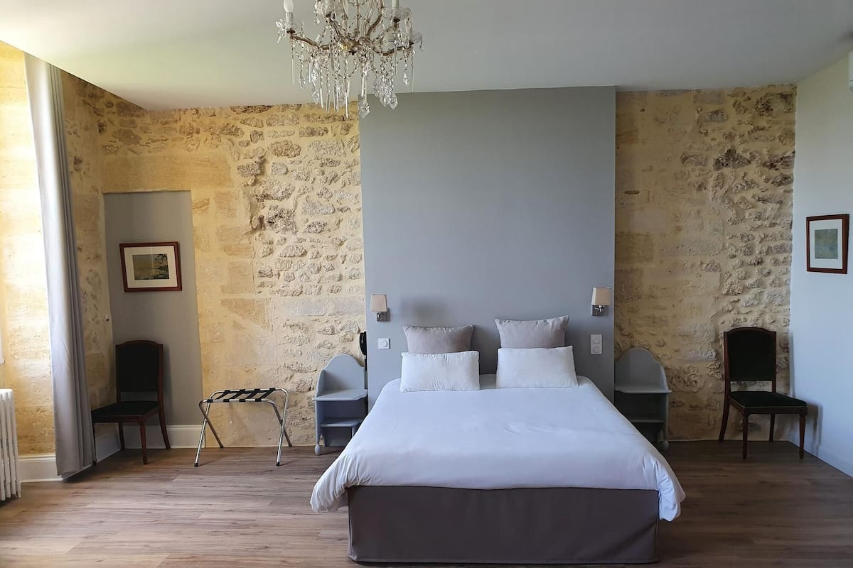 A bedroom with a large bed, two bedside tables, a chandelier, stone walls, and a radiator.