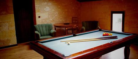 A pool table in a room with wooden floors and walls, a green armchair, and a small round table with a chair.