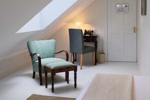 A room with a sloped ceiling, a wooden chair, a cushioned chair, a desk with a lamp, and a door with a light switch. - Chapter House Boutique Hotel (Franschhoek)
