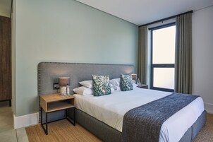 Un dormitorio ordenado con una cama grande, una mesita de noche con lámpara y una ventana con cortinas.