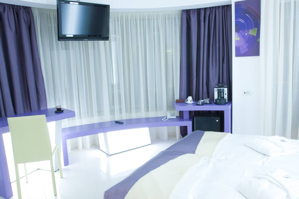 A modern hotel room with a bed, a desk, a chair, a TV, and a coffee machine.