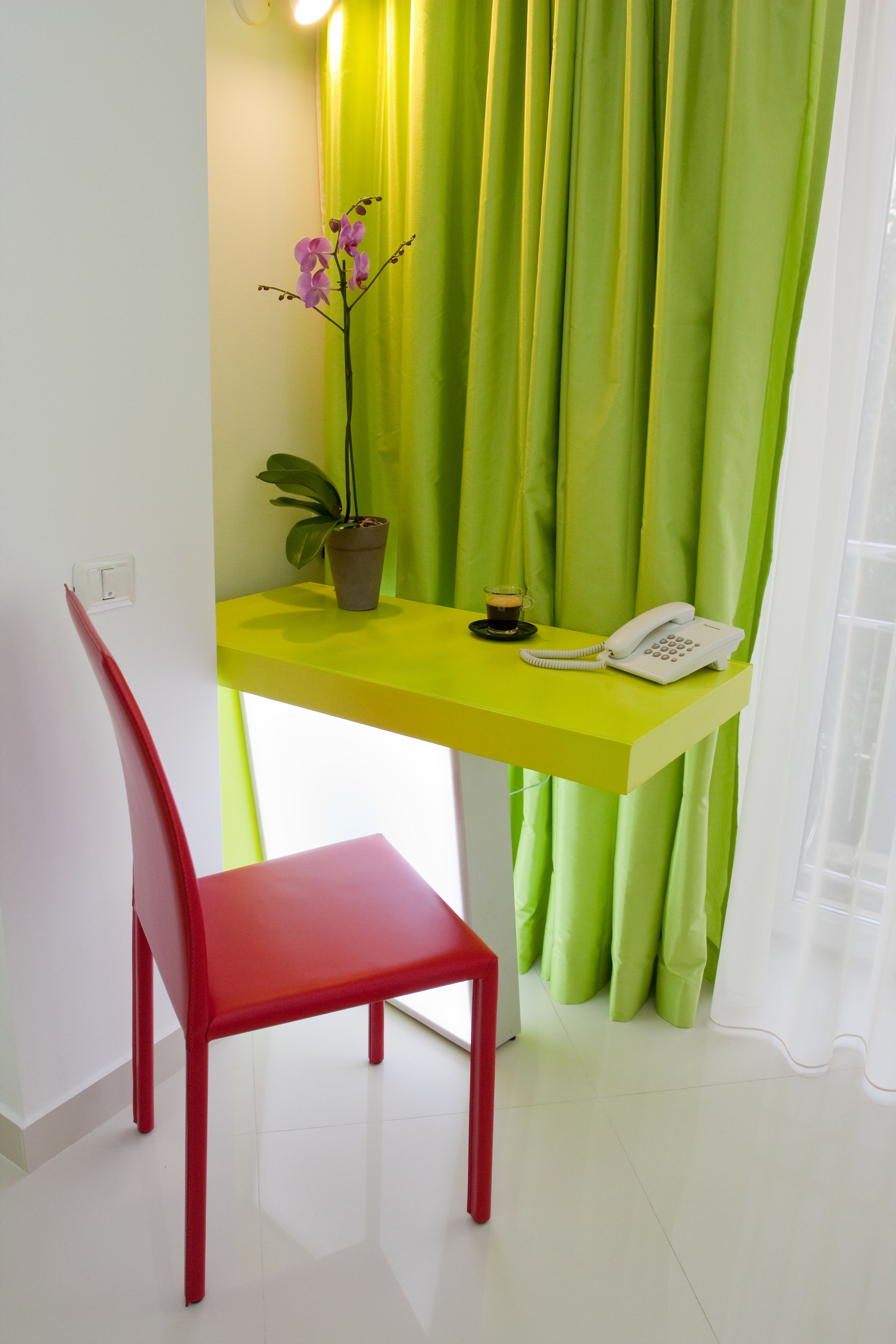 A bright room with a yellow desk, a red chair, a potted plant, a white phone, and a green curtain.
