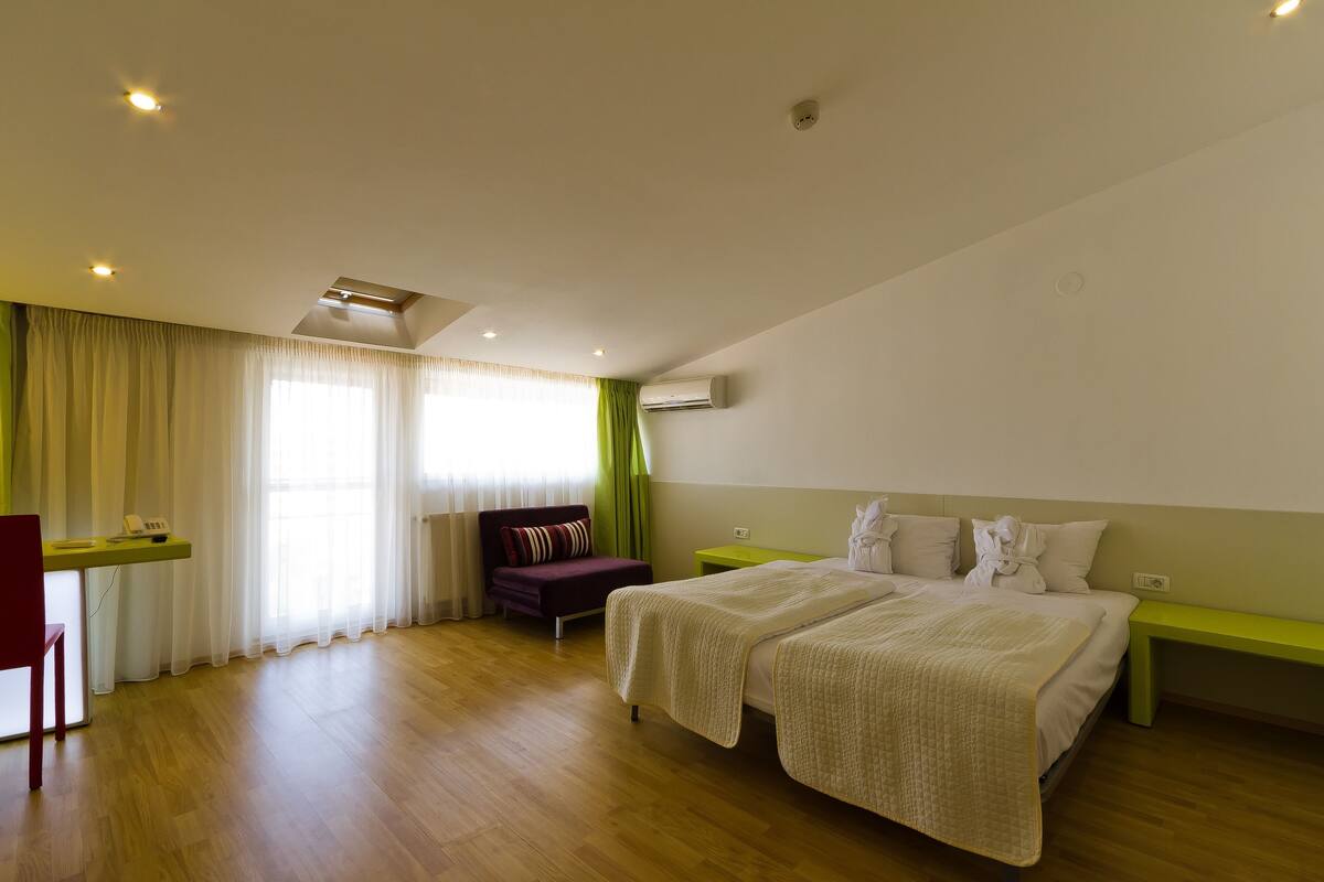 A hotel room with a wooden floor, two beds with white bedding, a desk with a chair, a green bench, and a window with sheer curtains.