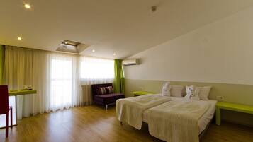 Une chambre d’hôtel avec un plancher en bois, deux lits avec du linge de lit blanc, un bureau avec une chaise, un banc vert et une fenêtre avec des rideaux légers.