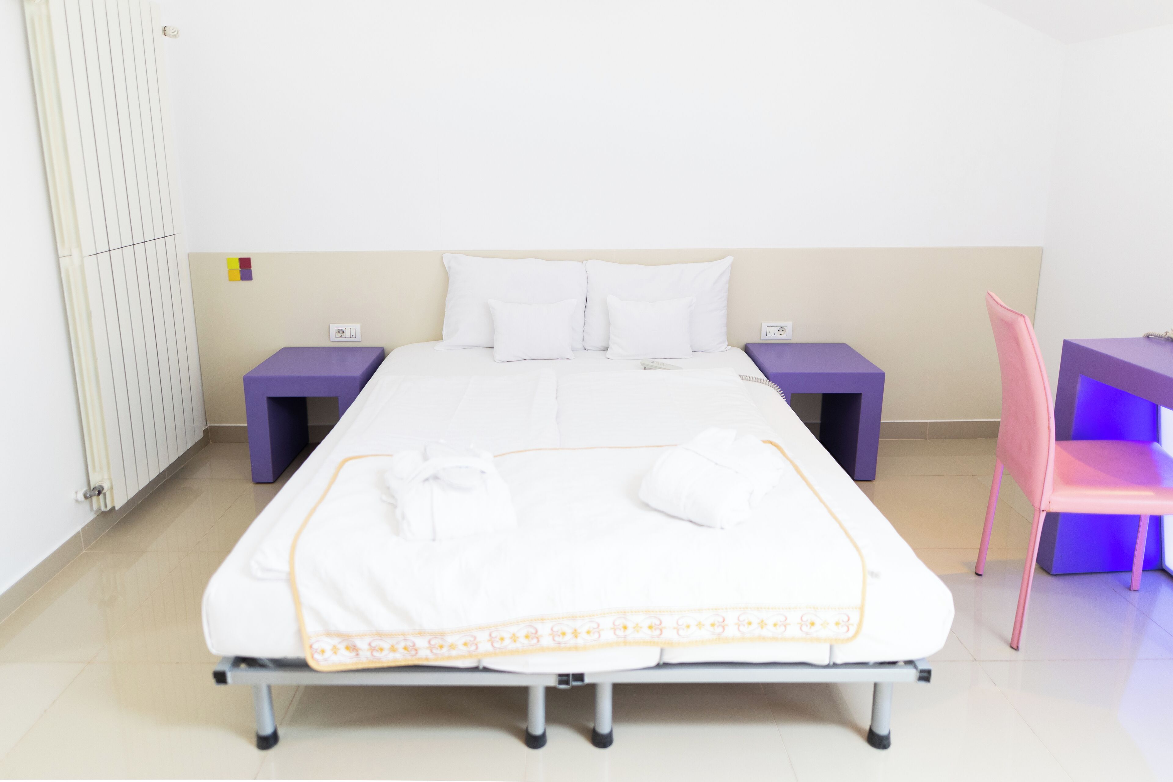 Una cama bien hecha con sábanas blancas y un borde decorativo, flanqueada por mesitas de noche moradas y una silla rosa.