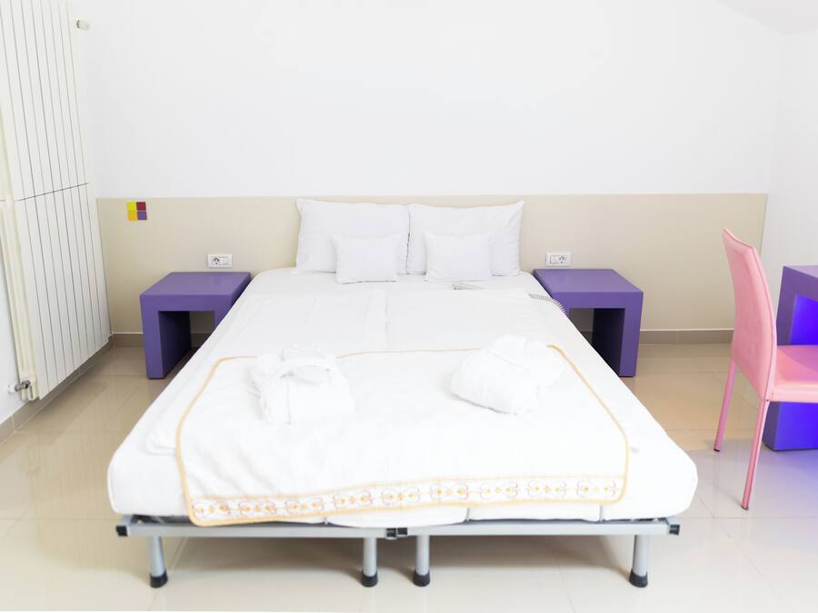 Una cama bien hecha con sábanas blancas y un borde decorativo, flanqueada por mesitas de noche moradas y una silla rosa.