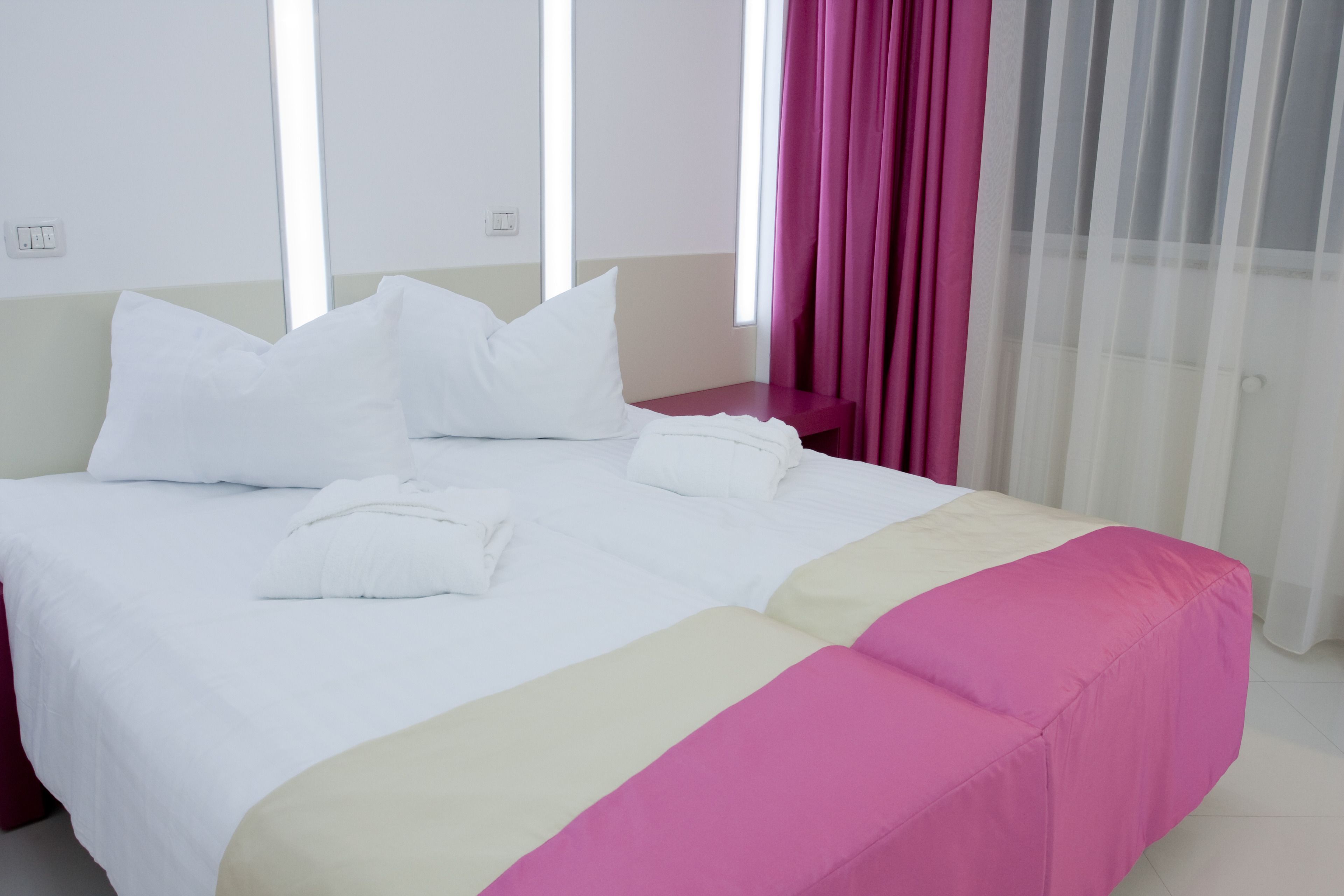 A neatly made bed with white linens and a pink accent, a bedside table, and a window with sheer curtains.