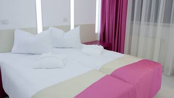 A neatly made bed with white linens and a pink accent, a bedside table, and a window with sheer curtains.