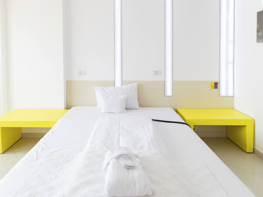 Ein ordentlich bezogenes Bett mit weißen Leinen, einem weißen Kopfkissen und einem gefalteten Handtuch auf einer weißen Matratze. Auf jeder Seite des Bettes befinden sich jeweils ein gelber Nachttisch.