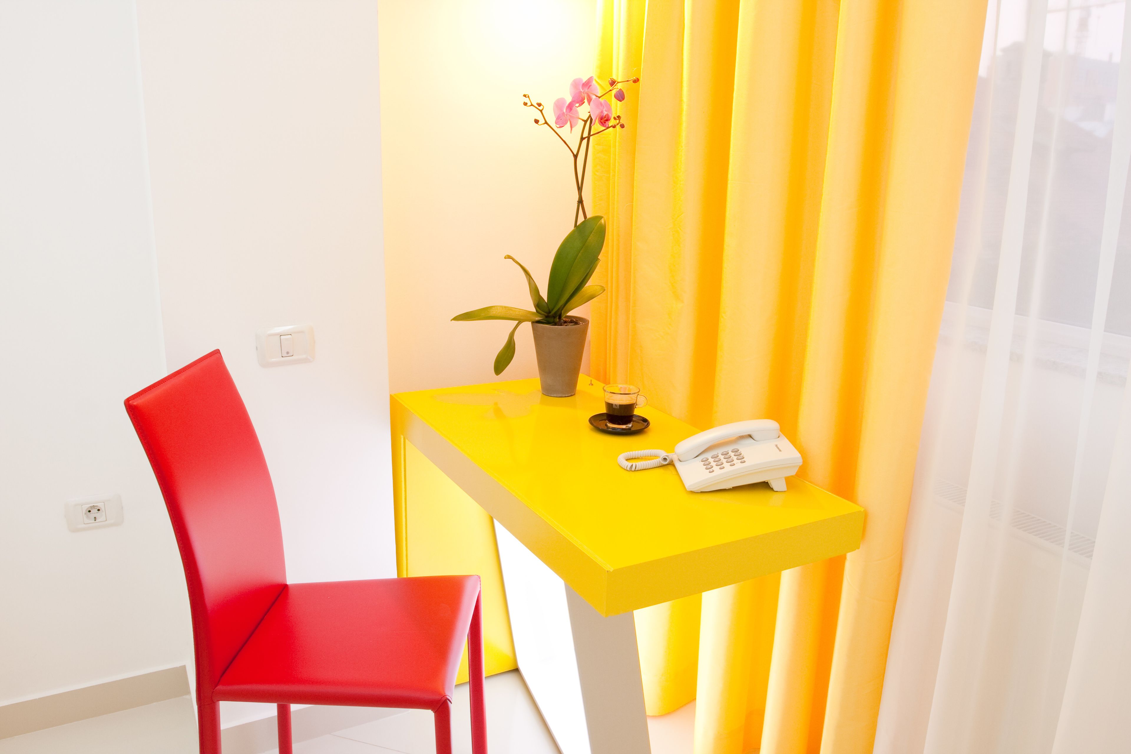 Ein helles Zimmer mit einem gelben Tisch, einem roten Stuhl, einer Topfpflanze und einem Telefon.