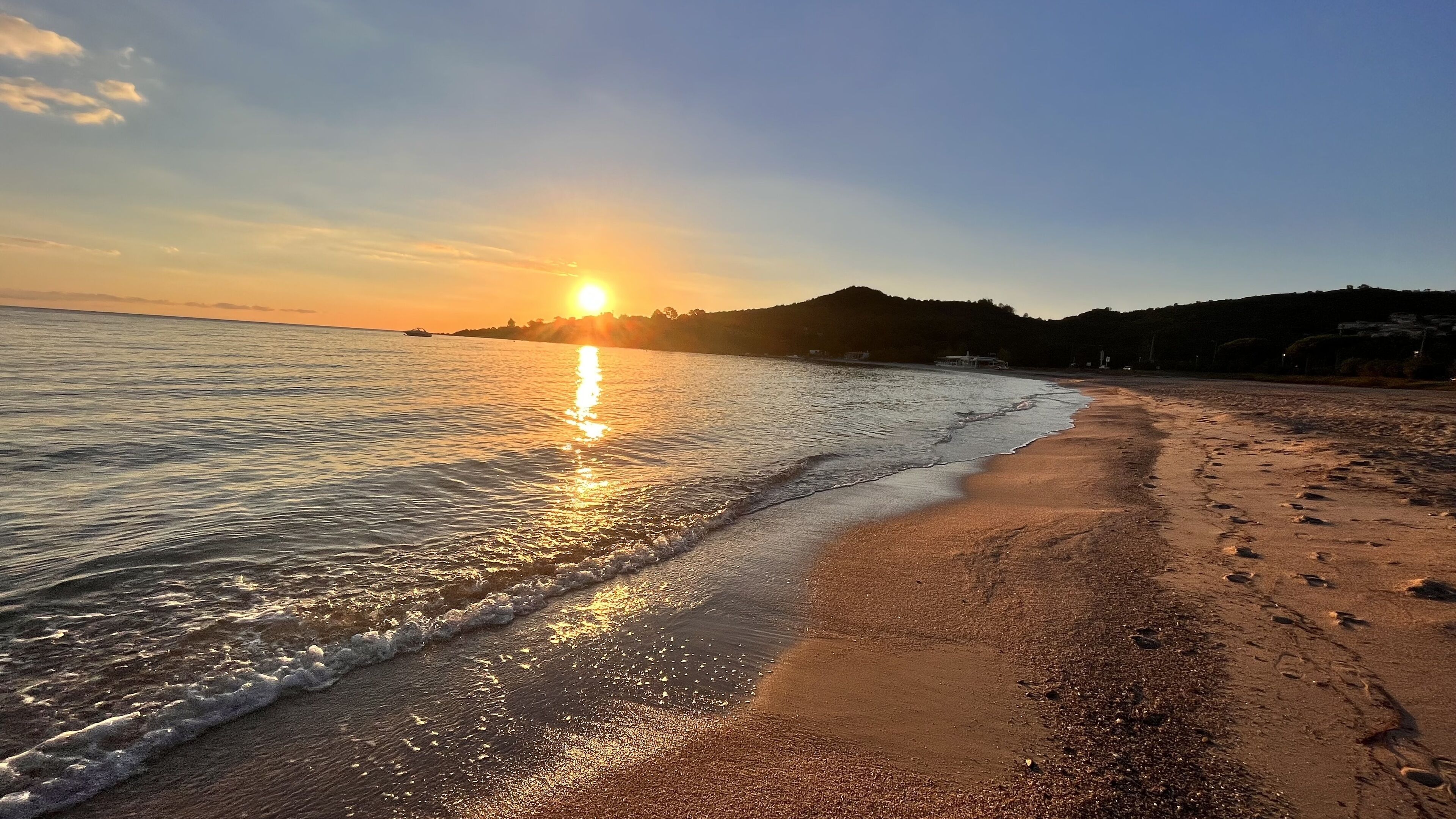 Ranta, josta avautuu näkymä auringonlaskuun veden yllä, hiekkainen rantaviiva ja kauempana kohoava kukkula.