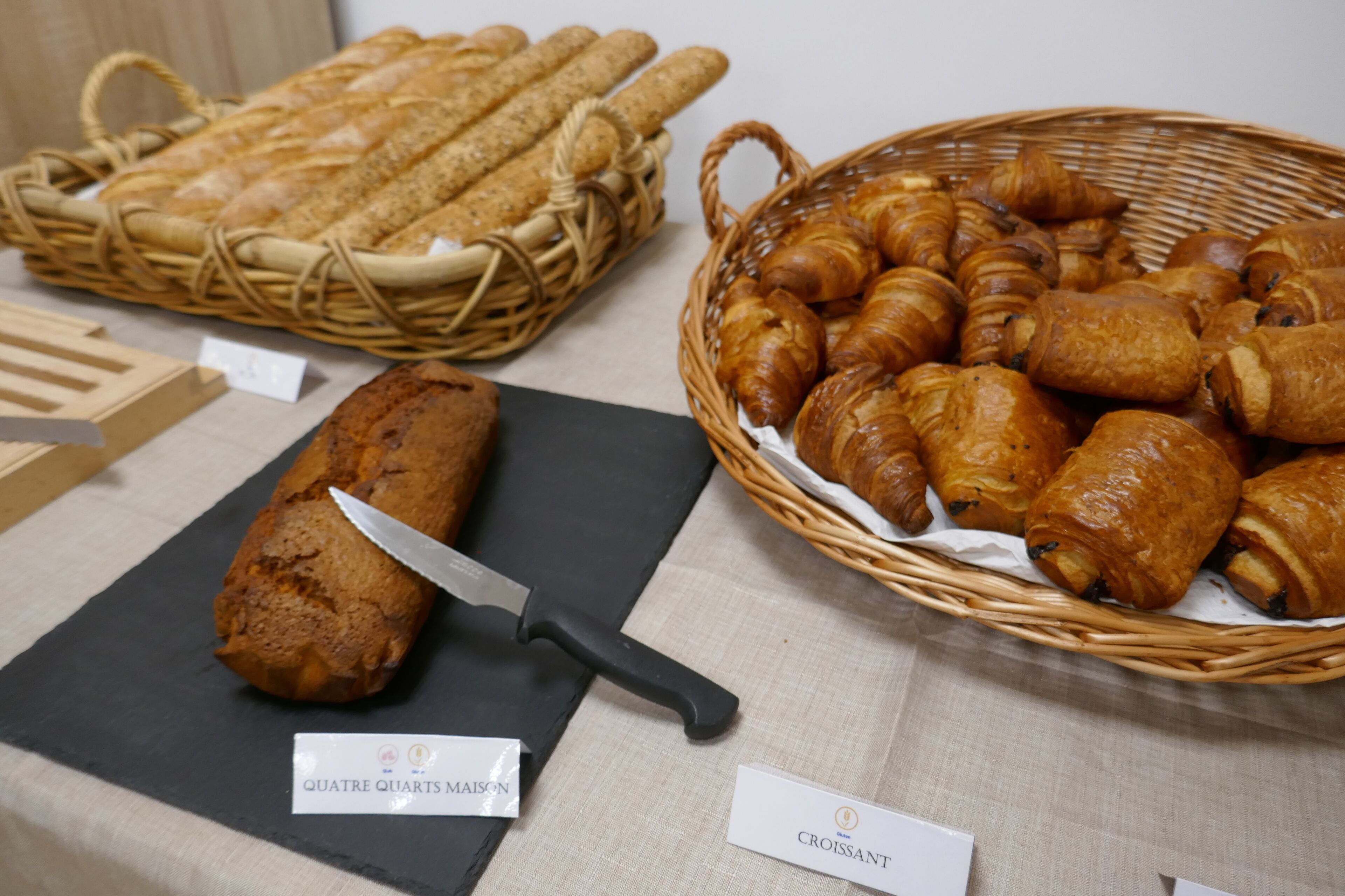 A display of baked goods including bread, croissants, and a loaf of cake with a knife on a black cutting board.
