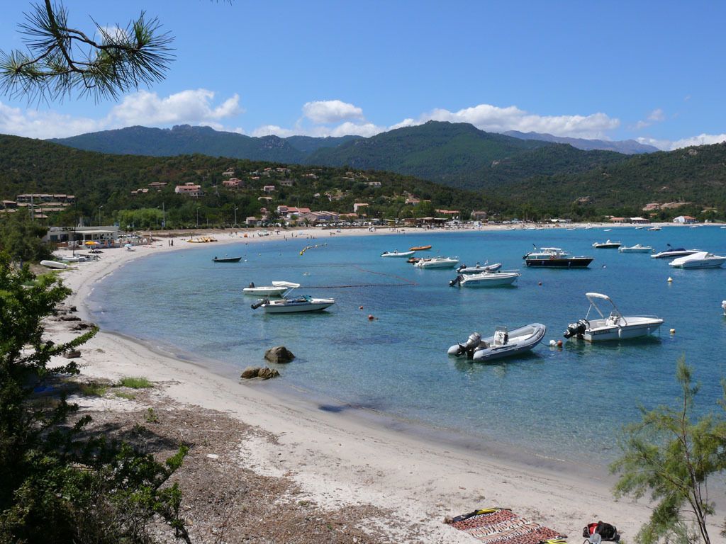 Une zone côtière où des bateaux sont amarrés près d’une plage de sable, le tout étant entouré de collines et de bâtiments.
