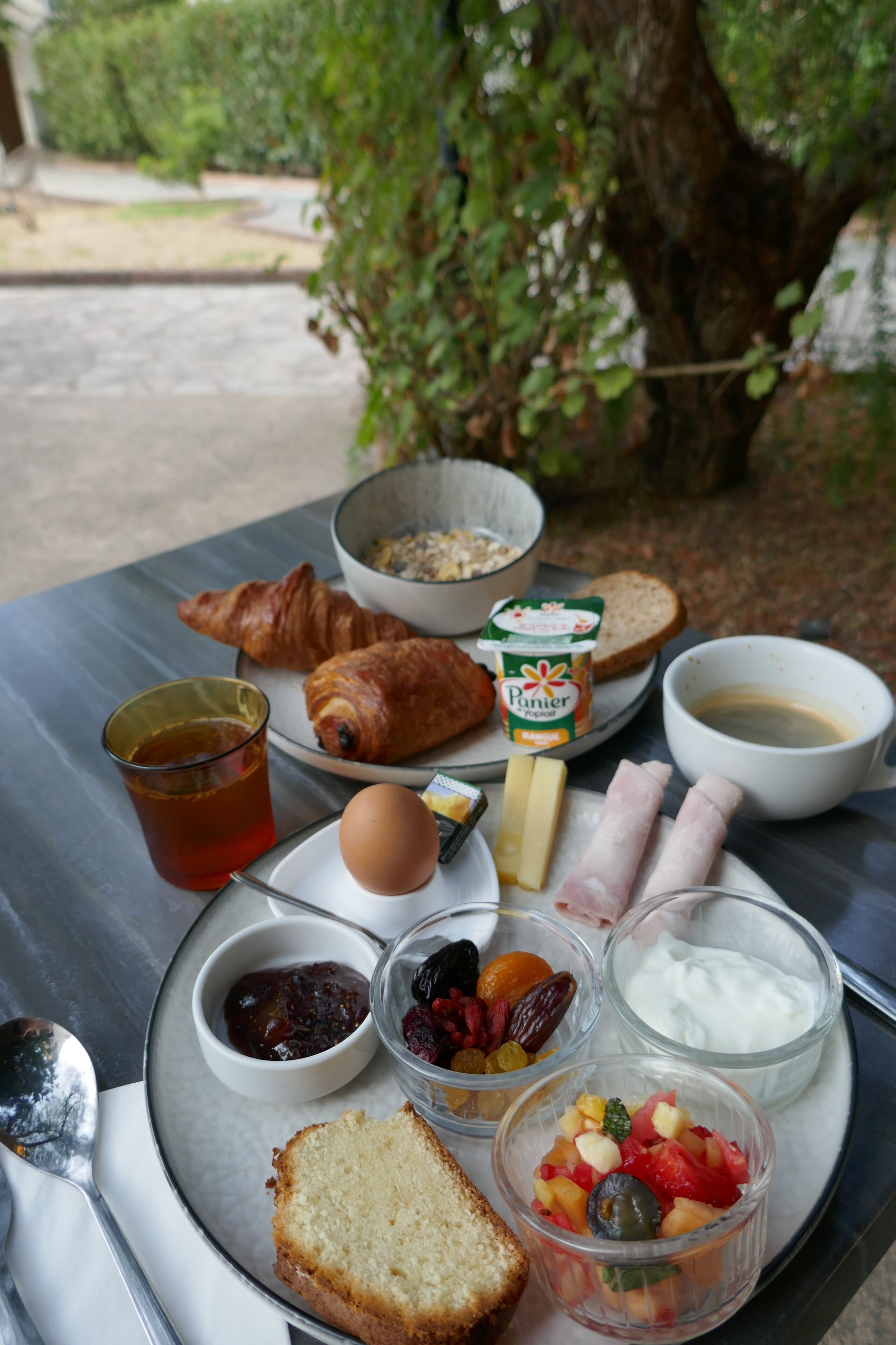 Ein Frühstückstisch mit Croissants, Brot, Käse, Schinken, Eiern, einer Schüssel Müsli, einer Tasse Tee sowie verschiedenen Obst- und Joghurtbechern.