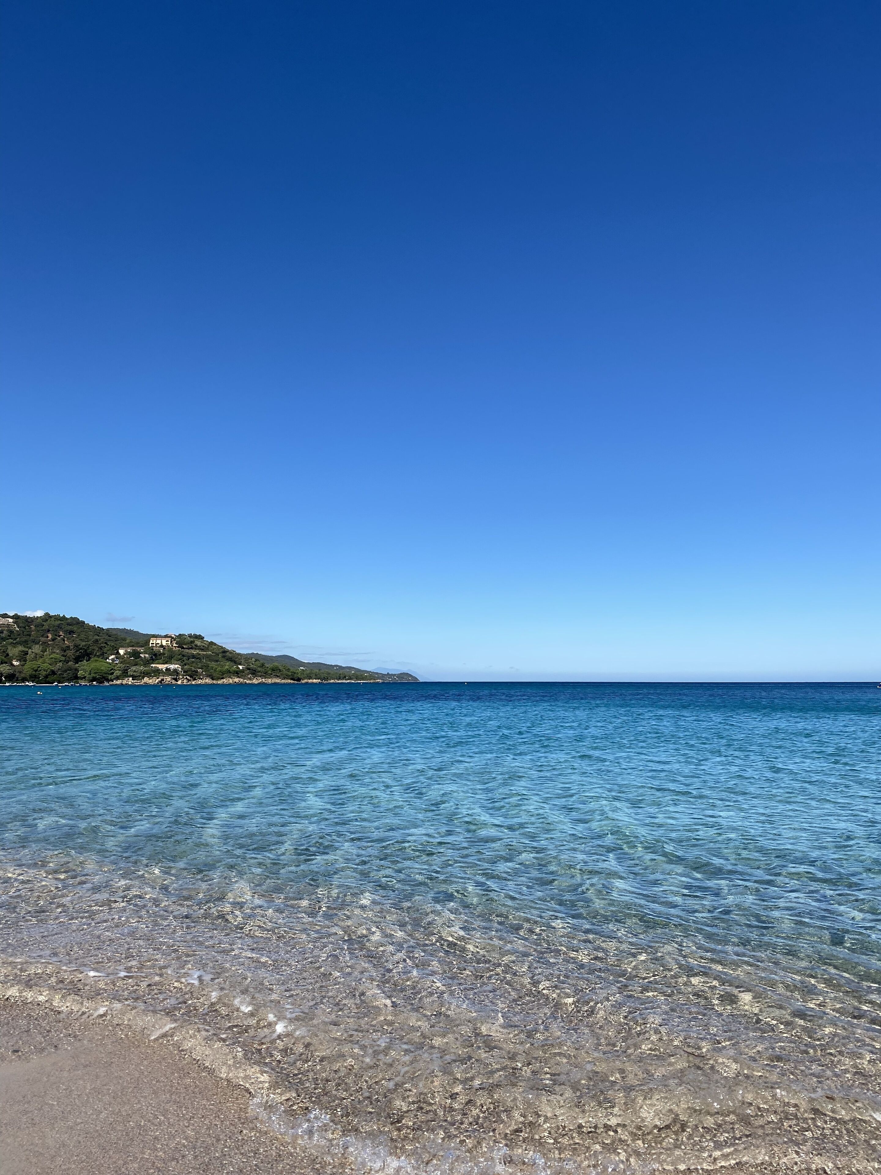 Vista despejada de una playa con aguas poco profundas, una costa lejana con edificios y un cielo azul claro.