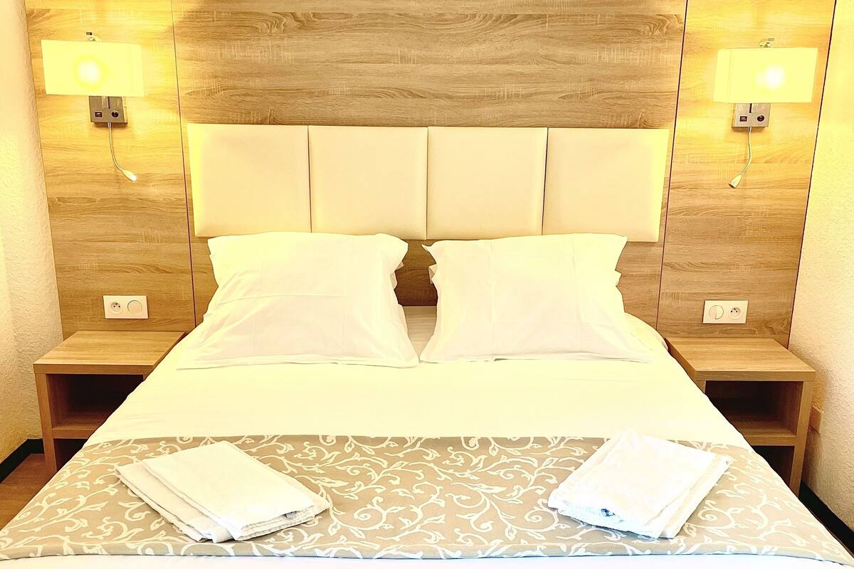 Una cama bien tendida con sábanas blancas y dos toallas dobladas, un cabecero de madera y lámparas de noche.