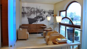 A modern lounge with a mural of a historic harbor scene, comfortable seating, and a view of the water through large windows. - Atrium Hotel (Elblag)