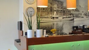 A modern reception desk with a green-lit lower section, a mural of a ship on the wall, and three wall clocks. - Atrium Hotel (Elblag)