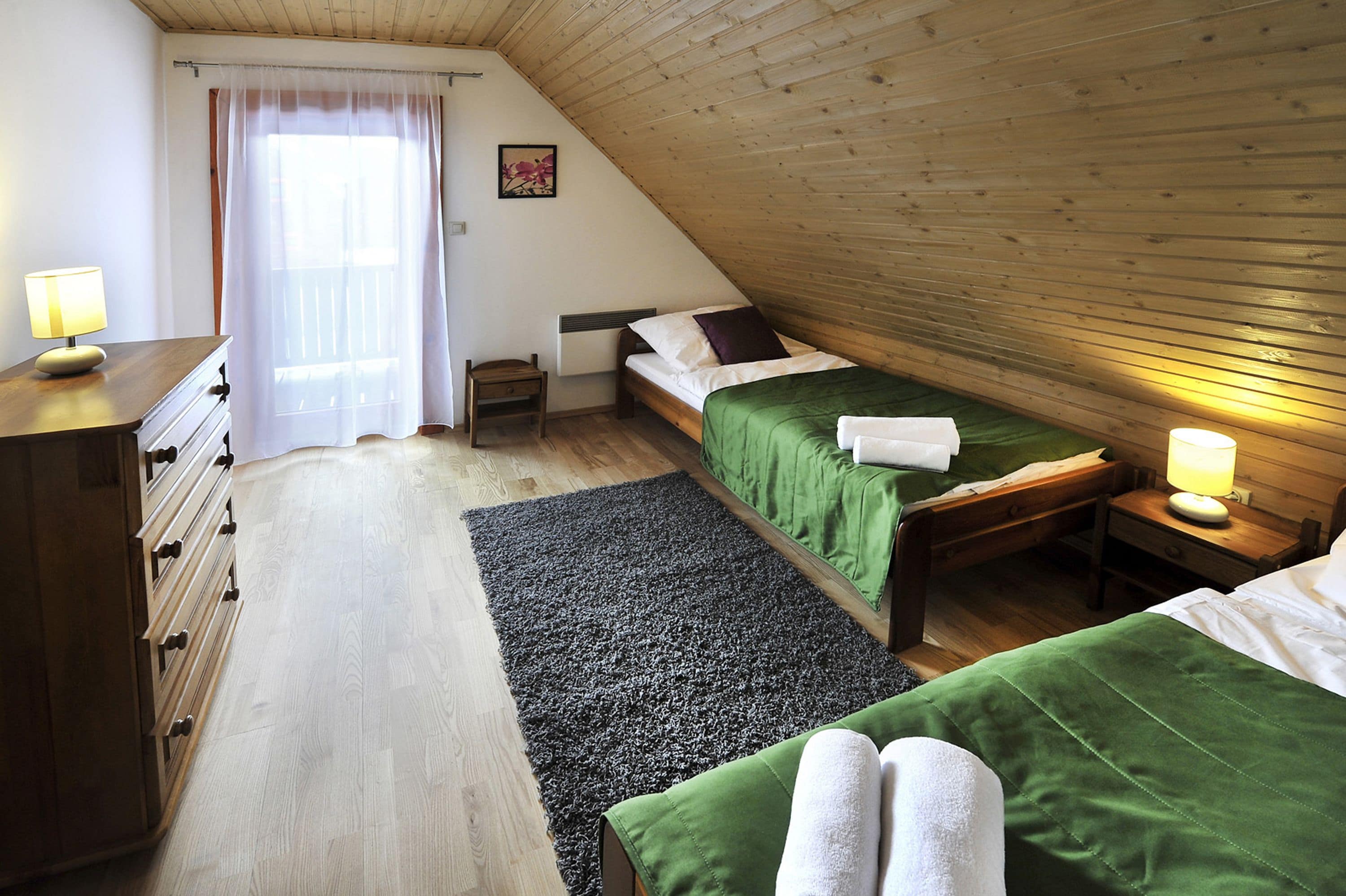 A cozy attic bedroom with a sloped ceiling, a wooden dresser, a bed with green bedding, and a nightstand with a lamp.