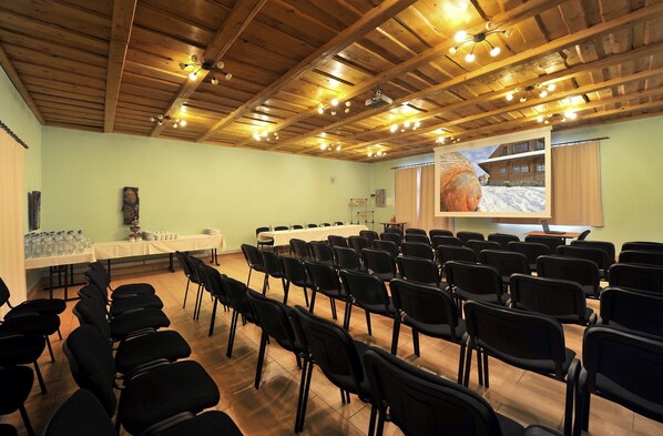 Un salón de conferencias con filas de sillas, una mesa larga y una pantalla proyectando un paisaje nevado.