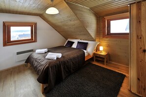 Une chambre avec un plafond en pente, un lit recouvert d’une couvre-lit marron foncé, deux lampes de chevet et une vue par la fenêtre.