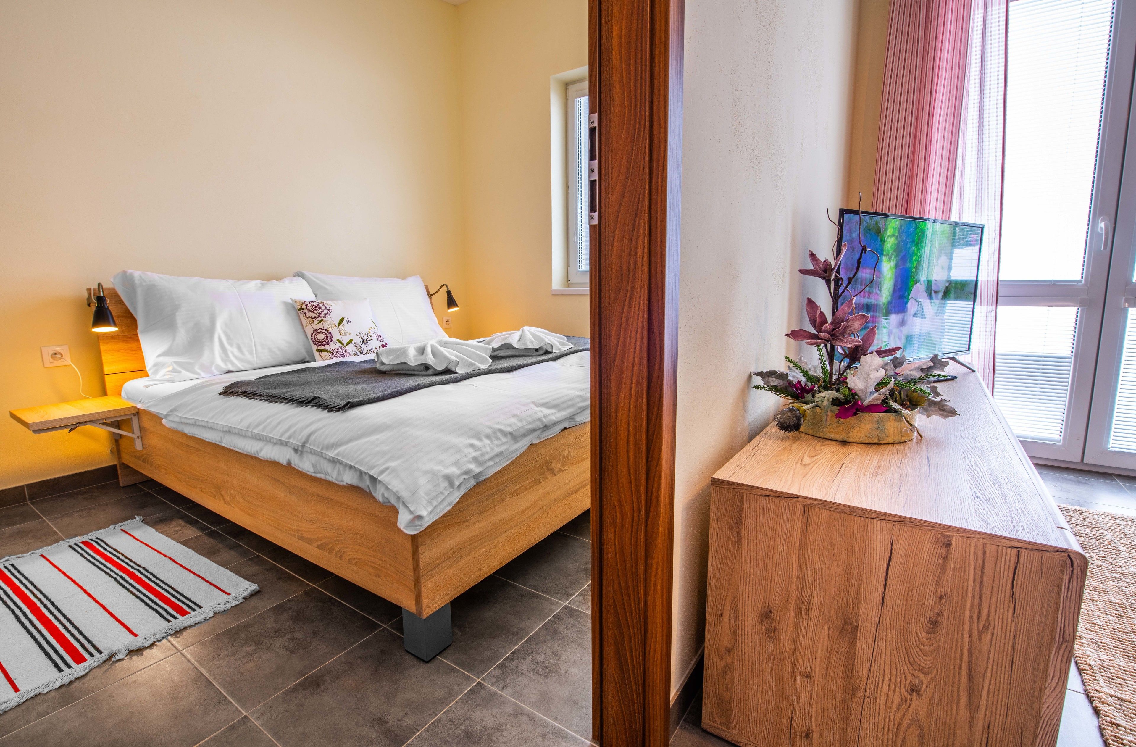 Un dormitorio con cama de madera, una mesita de noche con lámpara, una ventana con persianas y vistas al exterior.