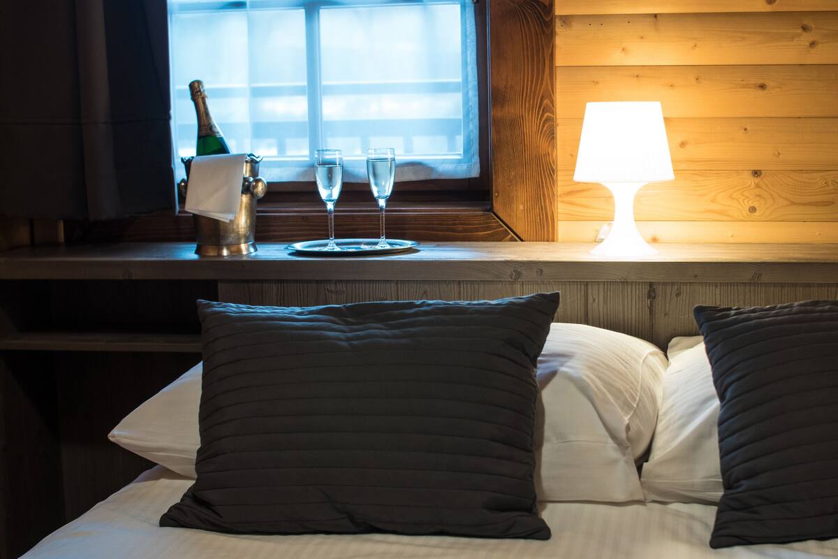Hotelkamer met houten hoofdeinde, nachtkastje met champagnefles, twee glazen en een lamp.
