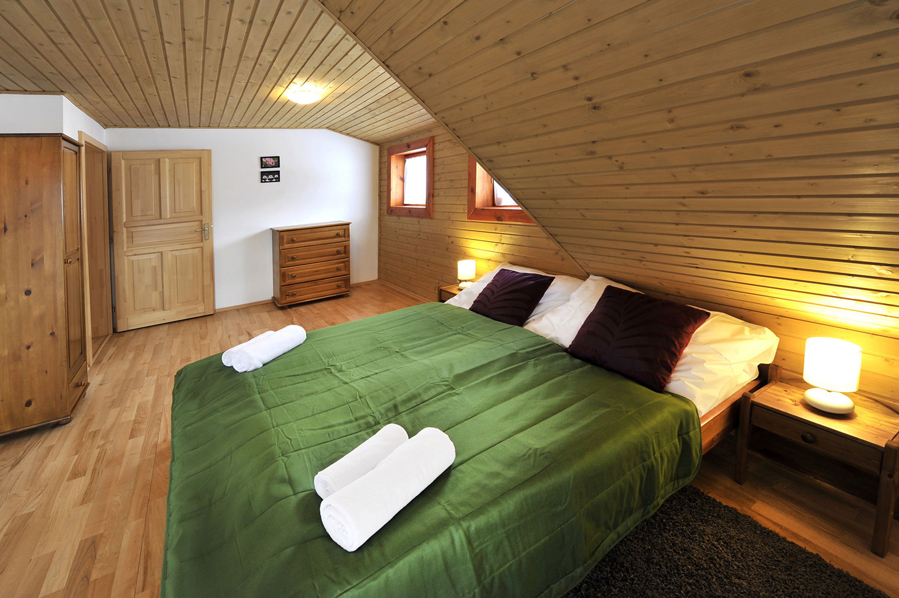 A bedroom with a wooden ceiling, a green bedspread, two white towels, a wooden nightstand with a lamp, and a wooden wardrobe.