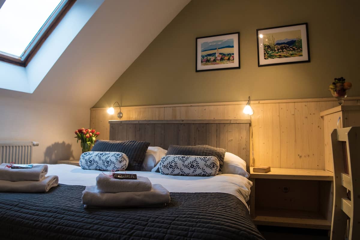 Een slaapkamer met een schuine plafond, een bed met donkere beddengoed, twee gestreepte kussens, een nachtkastje met een lamp en twee ingelijste foto's aan de muur.