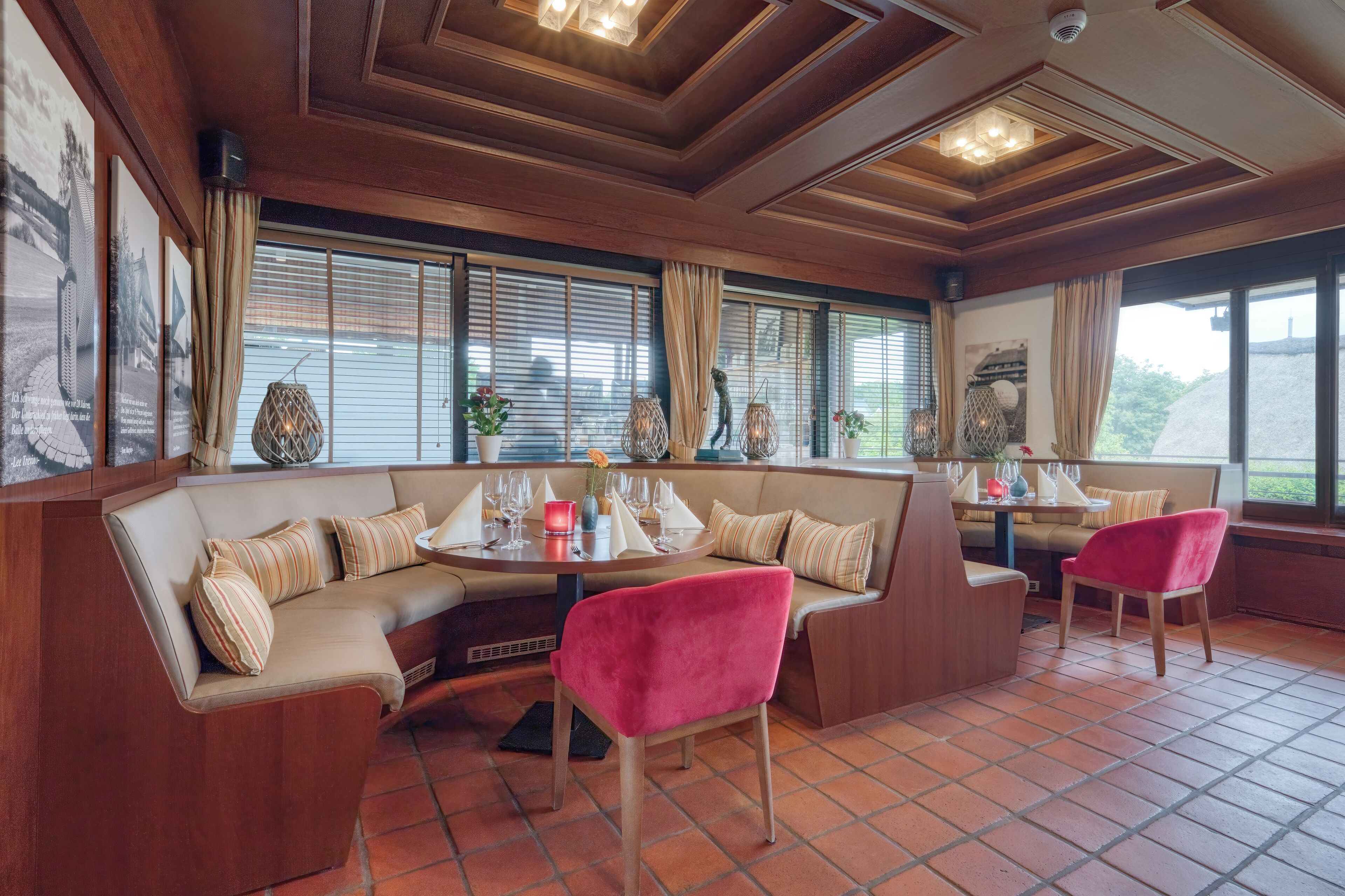 Un rincón comedor acogedor con un banco de madera y asientos acolchados, una mesa redonda con un jarrón y copas de vino, y ventanas amplias con persianas.