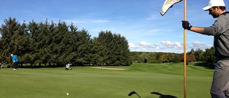 A golf course with a flagstick, a golfer in a blue jacket, and another golfer in the distance.