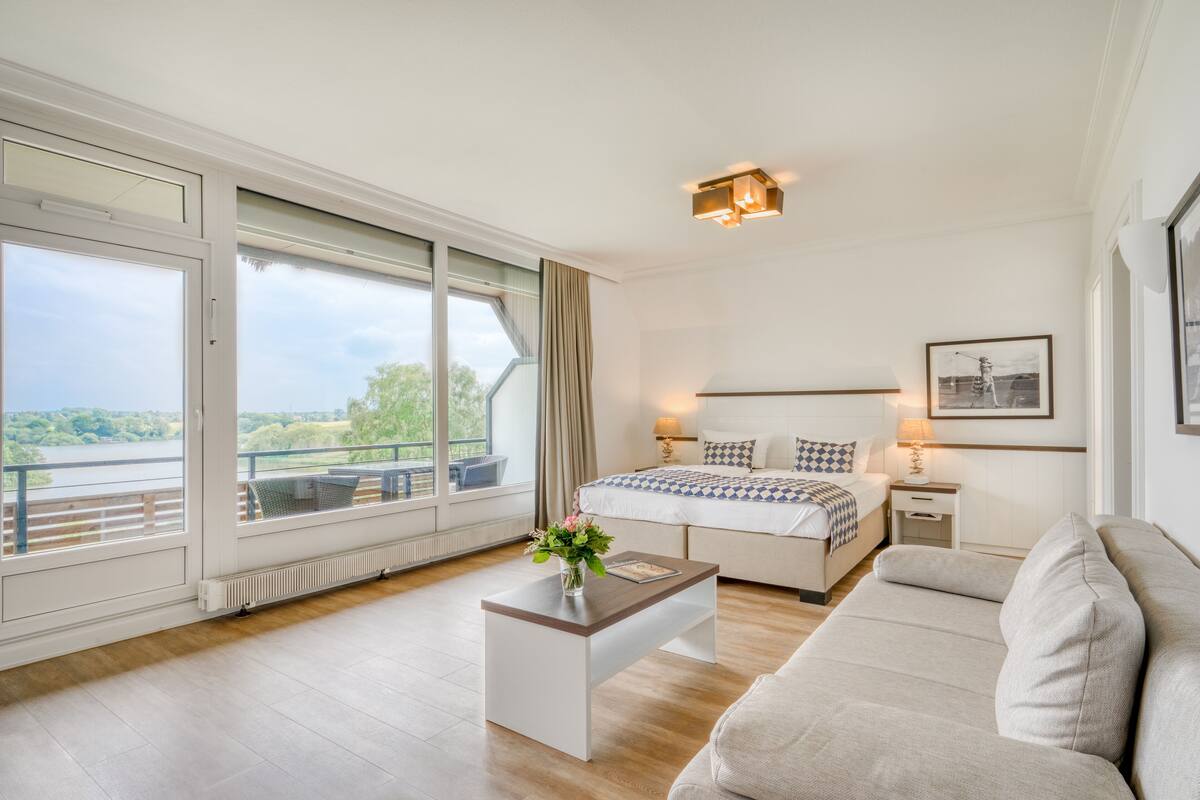 A modern bedroom with a large window, a bed with a patterned bedspread, a bedside table with a lamp, and a view of greenery outside.