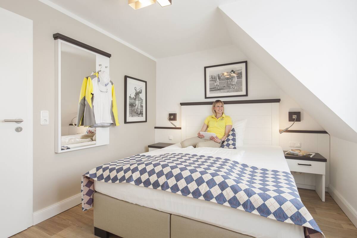 A bedroom with a bed, a nightstand, a mirror, and a wall-mounted shelf with a jacket and a framed picture.