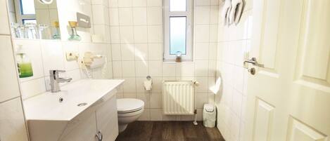 A small, clean bathroom with a toilet, sink, and mirror. There is a window above the toilet and a radiator below it.