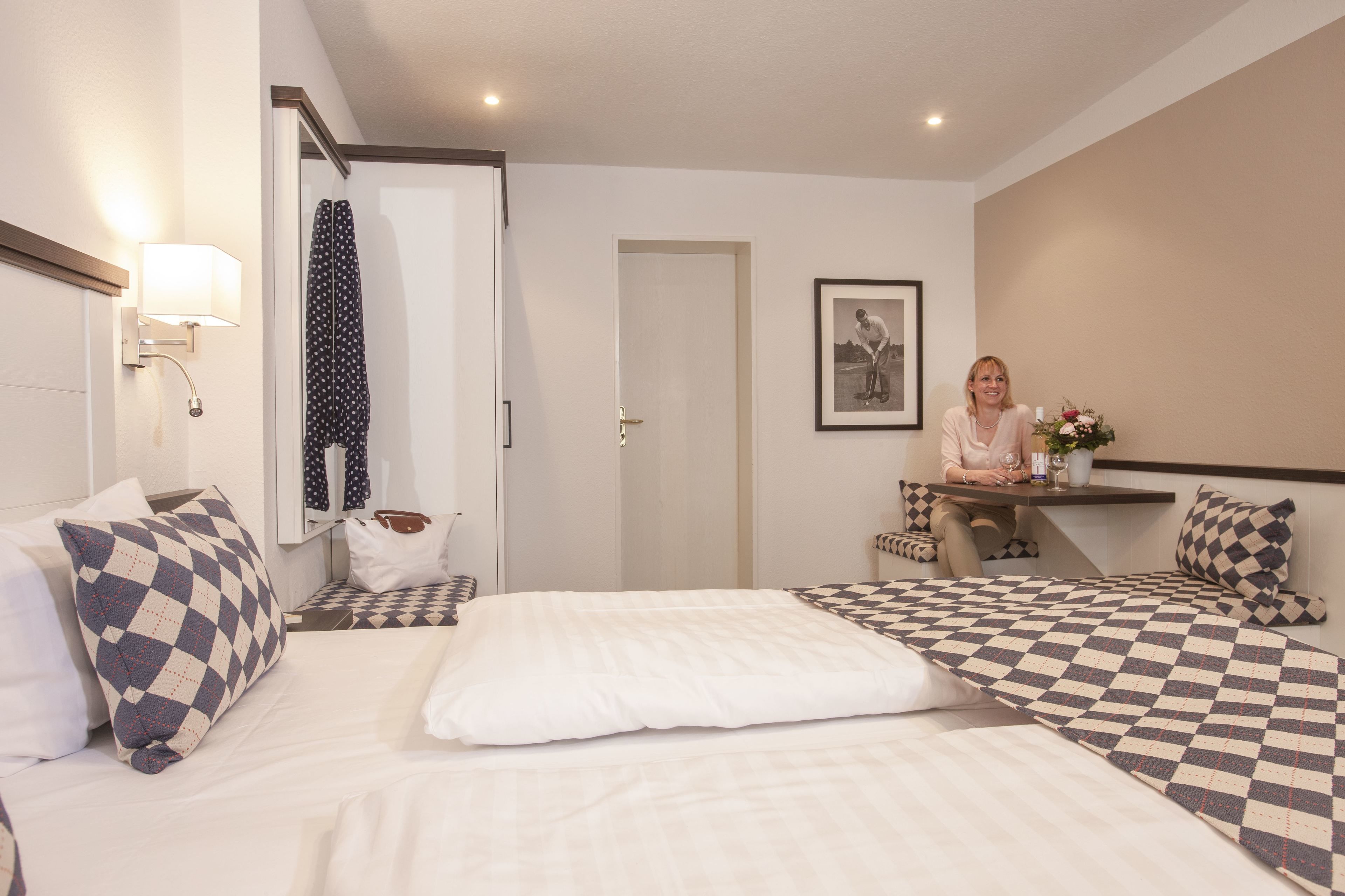 Ein Hotelzimmer mit Bett, Schreibtisch und Stuhl. Es gibt ein Fenster mit Vorhängen, eine Wandlampe und ein gerahmtes Bild an der Wand.