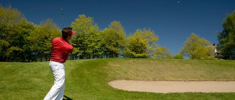 A person in a red shirt and white pants on a golf course with a clear sky and trees in the background.