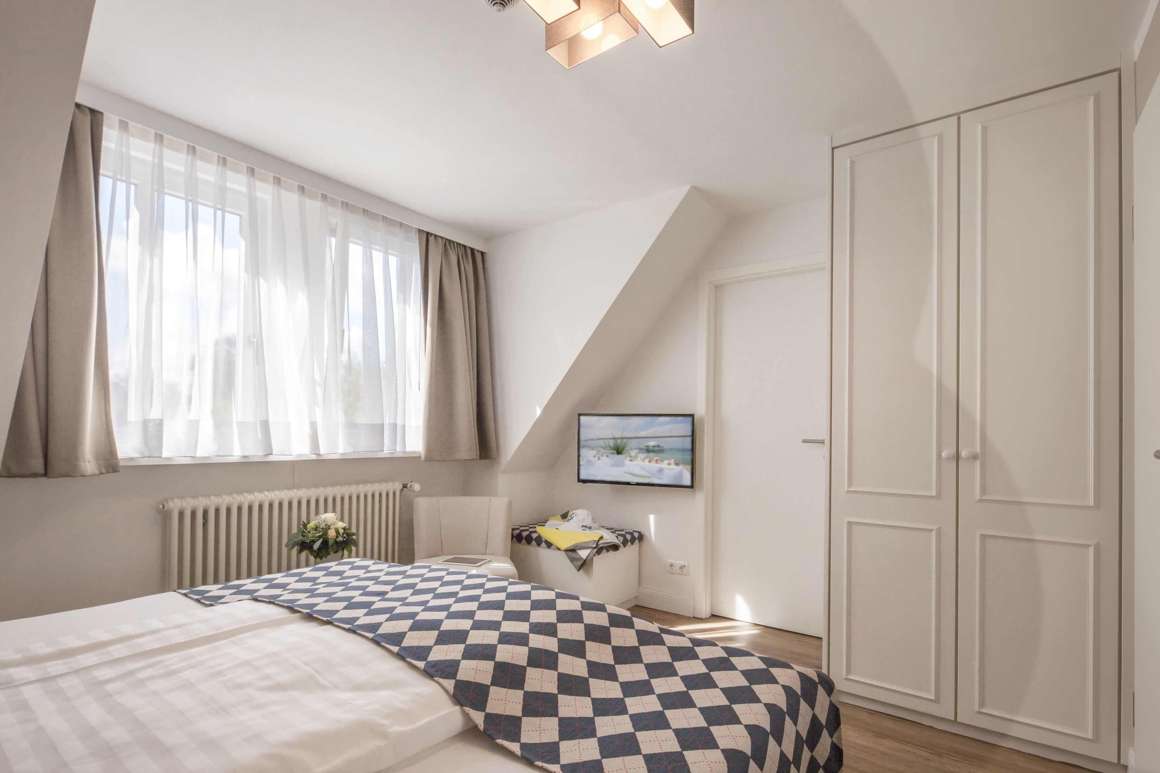 Ein Schlafzimmer mit Bett, Heizkörper, einem Fenster mit Vorhängen, einem Fernseher und einem Kleiderschrank.
