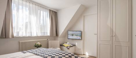 A bedroom with a bed, a radiator, a window with curtains, a TV, and a wardrobe.
