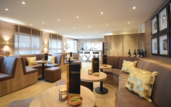 A modern lounge area with sofas, round tables, and a bar area in the background. - Strandgrün Golf- & Spa Resort (Timmendorfer Strand)