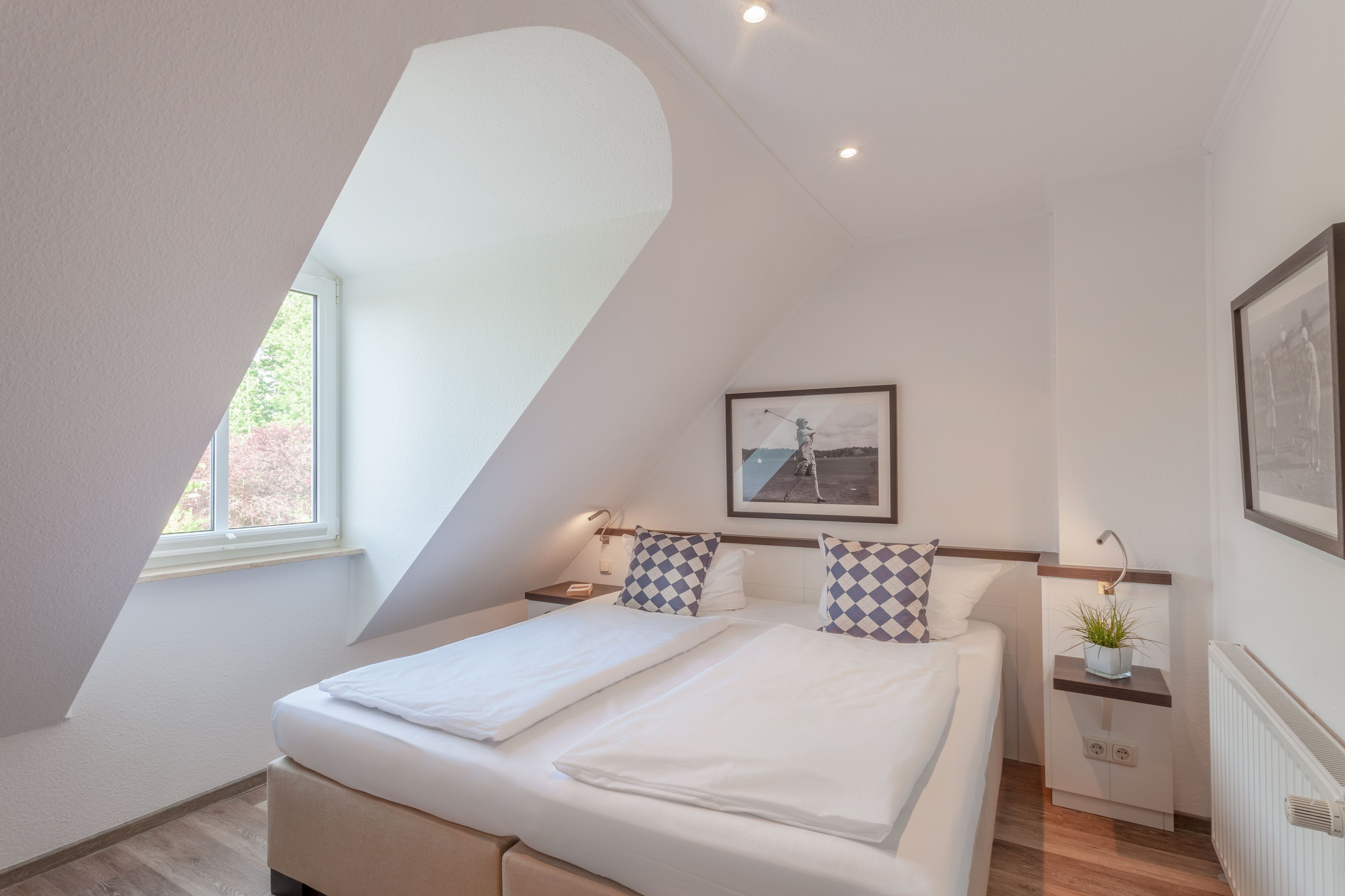 A bedroom with a sloped ceiling, a bed with white bedding, a nightstand, a wall-mounted shelf with a plant, and a framed picture above the bed.