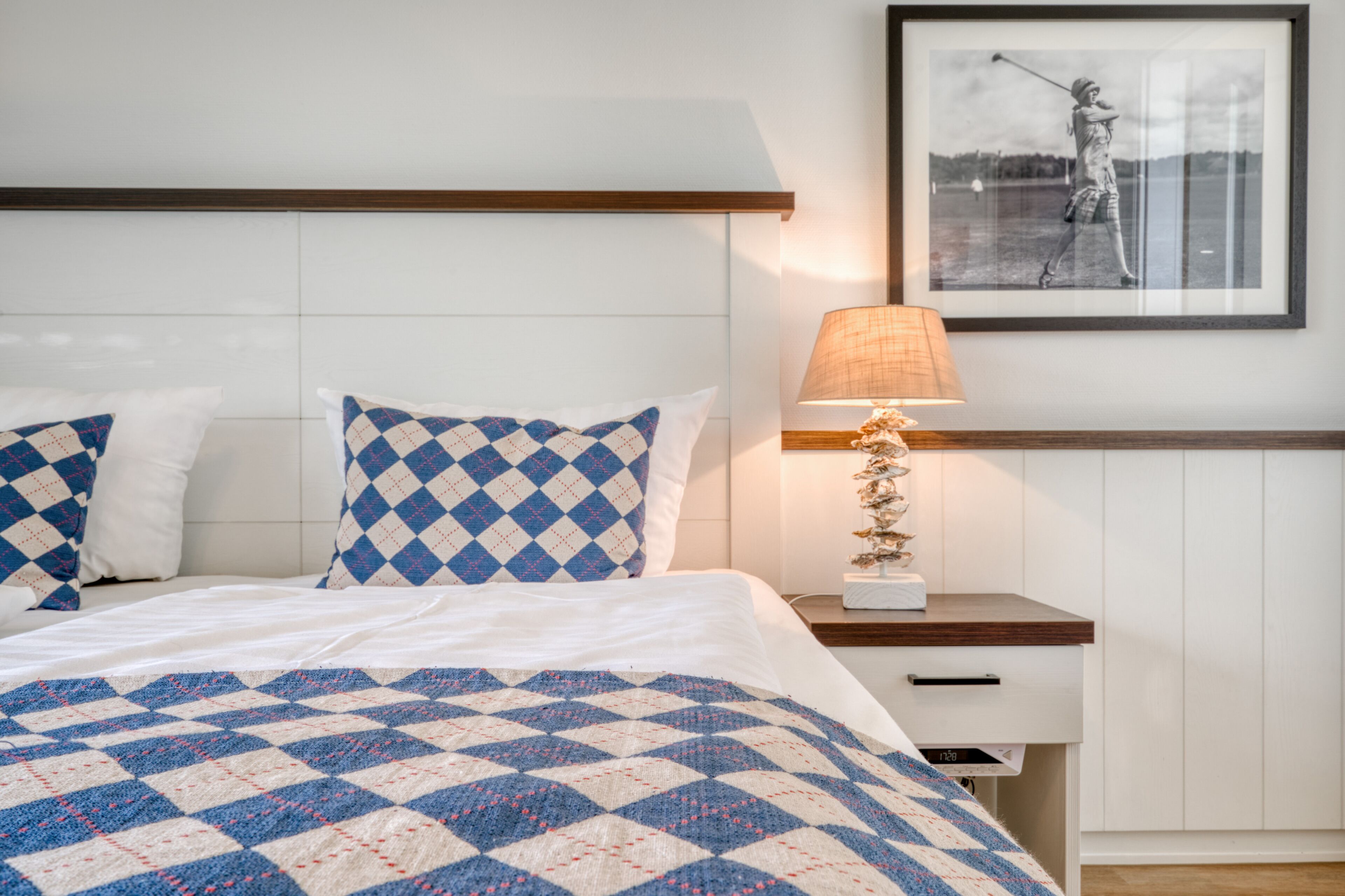 A neatly made bed with a blue and white checkered blanket, a bedside lamp, and a framed photograph on the wall.