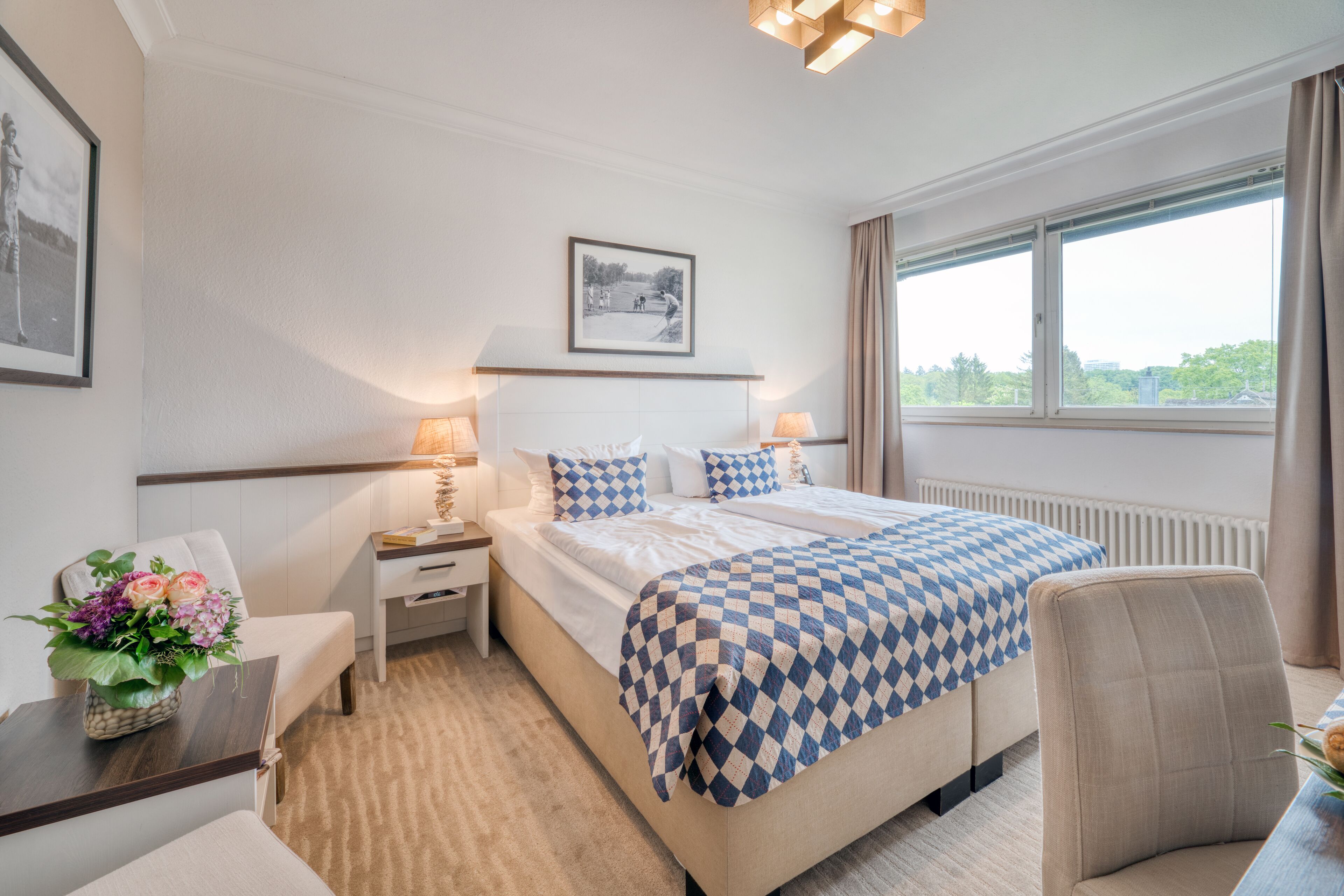 A hotel room with a large bed, two bedside tables with lamps, a chair, a desk, and a window with a view of greenery.
