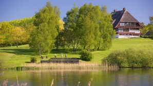 A house with a thatched roof, surrounded by a lush green landscape and a pond. - Strandgrün Golf- & Spa Resort (Timmendorfer Strand)