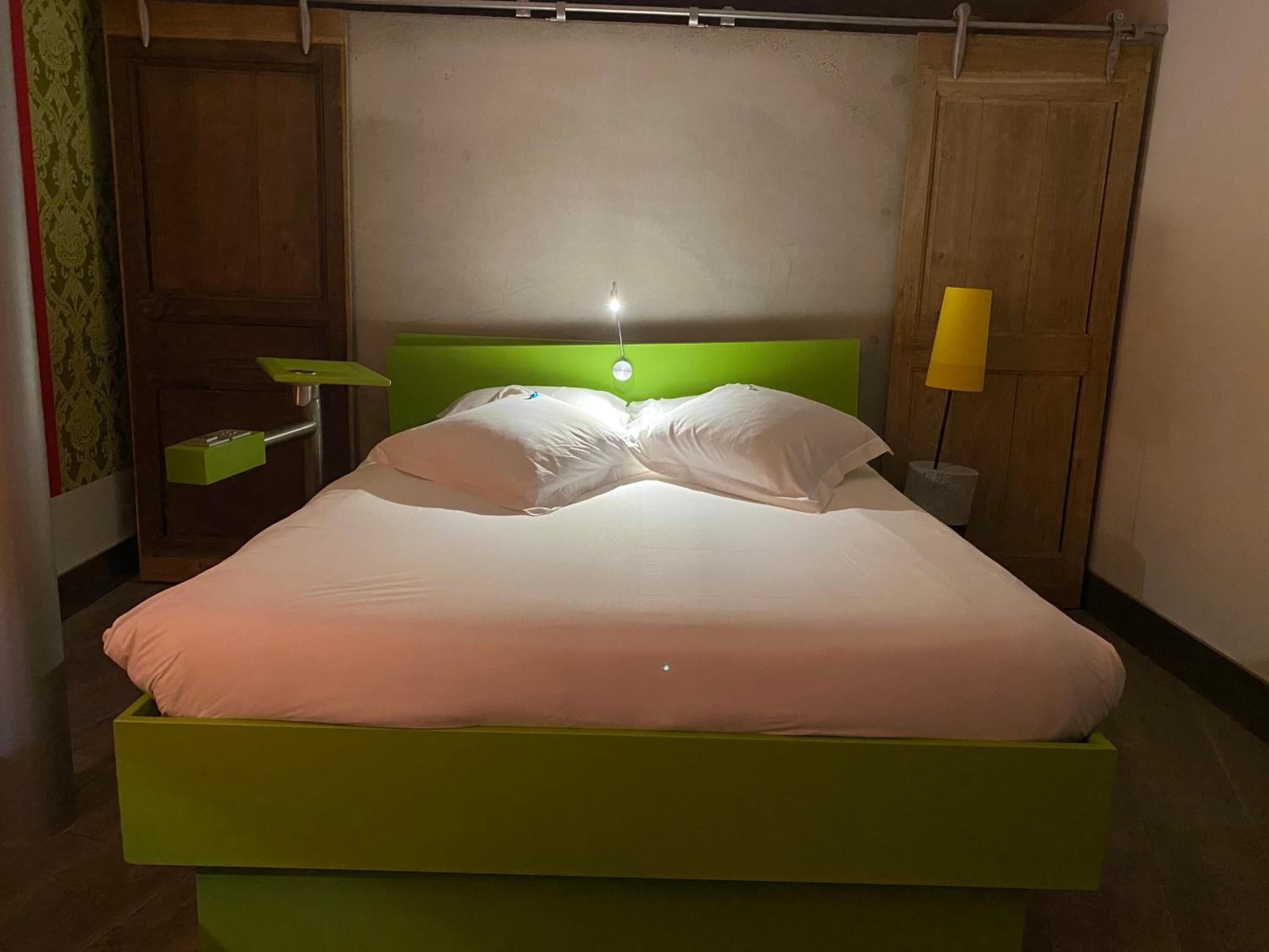 Una cama con cabecera verde y ropa de cama blanca, una mesita de noche con lámpara, y un armario de madera.
