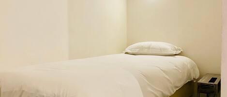 A single bed with a white mattress and pillows, a bedside table, and a wall-mounted light fixture.
